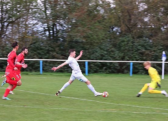 Ein Fußballspiel ist auf einem Feld im Gange. Drei Spieler verfolgen den Ball, und einer steht kurz davor, ihn zu schießen. Der Spieler in Weiß ist in der Luft, während die anderen in Bewegung sind. Eine weiße Linie ist auf dem Feld gezogen.