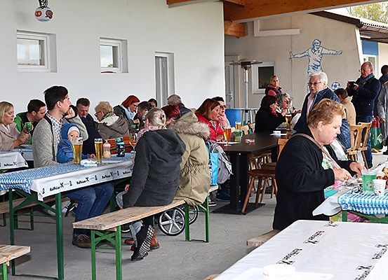 Eine Gruppe von Menschen, die an Tischen in einem überdachten Außenbereich sitzen, einige trinken Bier, eine Person im Rollstuhl und eine Wandmalerei an der Wand.