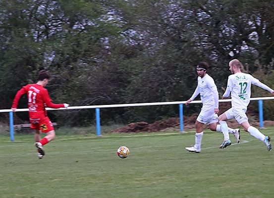 Drei Fußballspieler rennen auf einem Feld. Der Spieler in Rot versucht, den Ball zu treten. Die beiden Spieler in Weiß versuchen, ihn zu blockieren. Der Ball liegt auf dem Boden. Dahinter befindet sich ein Zaun mit einem blauen Pfosten. Dahinter stehen Bäume.