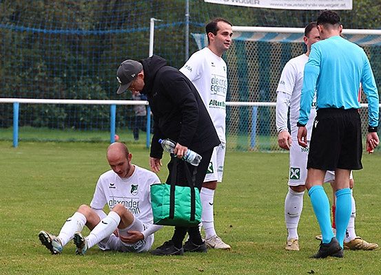 Ein Fußballspieler sitzt auf dem Boden, möglicherweise verletzt, während ein Trainer sich um ihn kümmert. In der Nähe stehen andere Spieler und ein Schiedsrichter auf dem Feld.