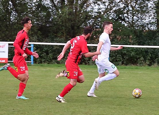Drei Fußballspieler sind auf einem Feld. Zwei tragen Rot, einer Weiß. Sie jagen einem Fußball hinterher. Dahinter steht eine weiße Barriere mit einem Schild.