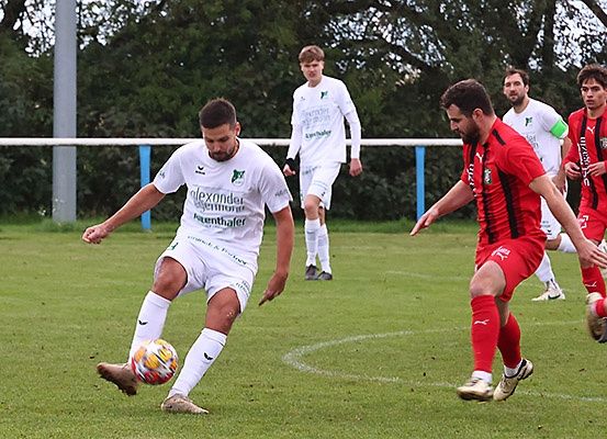 Fußballspieler auf einem Feld, einer in Weiß kickt den Ball, während zwei andere in Rot versuchen, ihn zu blocken. Bäume und ein Zaun im Hintergrund.