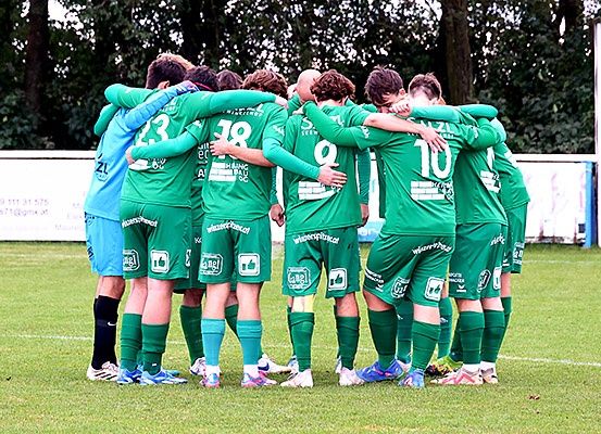 Eine Fußballmannschaft in grünen Trikots versammelt sich auf einem Feld mit Nummern auf dem Rücken, umgeben von Bäumen.