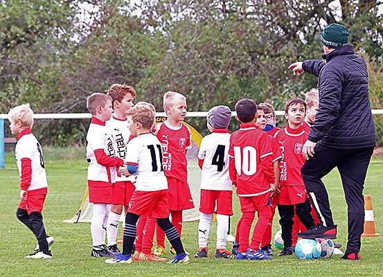 Ein Trainer in einer schwarzen Jacke und einer grünen Mütze instruiert eine Gruppe junger Fußballspieler in roten und weißen Trikots. Die Kinder stehen in einer Reihe auf einem Rasenfeld mit Fußballbällen und einem Verkehrskegel in der Nähe.