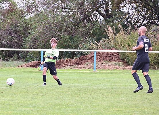 Ein Fußballspieler in einem grünen Trikot ist kurz davor, den Ball zu schießen, während ein anderer Spieler in einem schwarzen Trikot auf den Ball zuläuft.