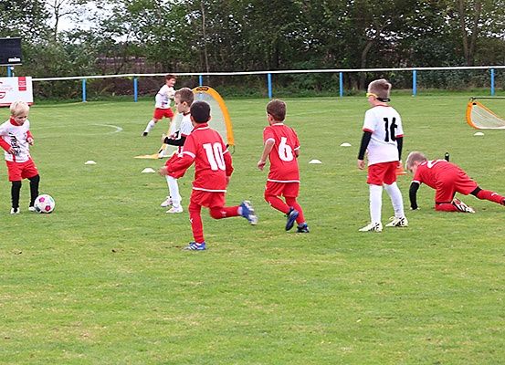 Eine Gruppe junger Jungen in roten Trikots mit den Nummern 6, 10 und 16 spielt Fußball auf einem Rasenfeld mit Toren und Verkehrskegeln.