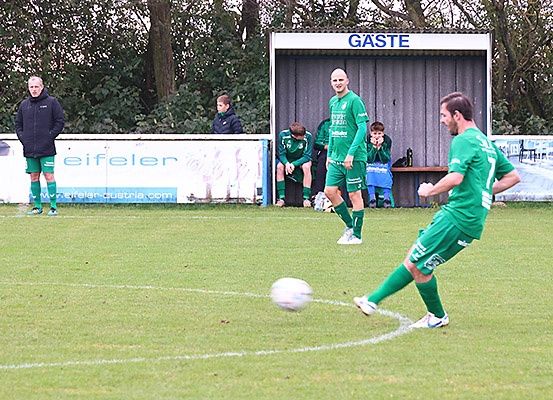 Fußballspieler in grünen Trikots auf einem Feld, einer kickt den Ball, ein anderer beobachtet, und zwei sitzen auf einer Bank mit einem Schild, das 'Gaste' steht.