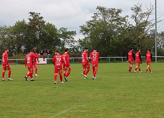 Eine Fußballmannschaft in roten Trikots läuft über ein Rasenfeld. Sie sind in einer Reihe aufgestellt und bereiten sich möglicherweise auf ein Spiel vor. Das Feld hat weiße Linien und ein Punkteanzeigebrett ist im Hintergrund zu sehen.