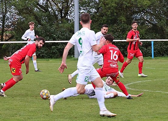 Fußballspieler in roten und weißen Trikots auf einem Feld, ein Spieler fällt, ein anderer tritt den Ball und ein Spieler im Hintergrund.