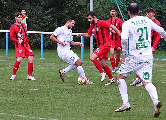 Ein Fußballspiel ist im Gange mit Spielern in roten und weißen Trikots. Ein Spieler in Weiß dribles den Ball, während andere in Rot versuchen, ihn zu blocken. Das Feld ist mit weißen Linien markiert.