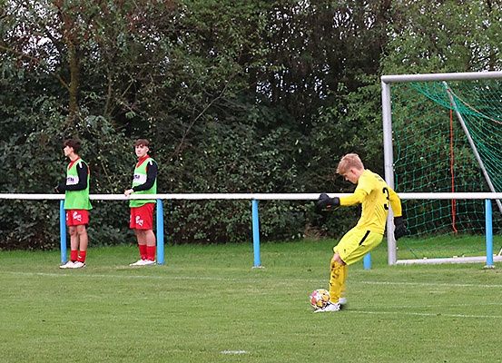 Drei Fußballspieler, einer tritt einen Ball, befinden sich auf einem Spielfeld mit einem Tor im Hintergrund.