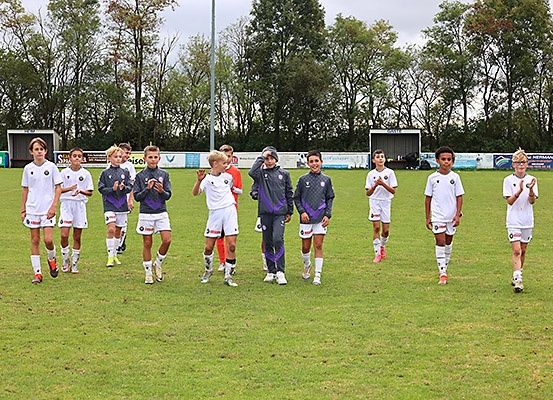 Eine Fußballmannschaft junger Spieler läuft auf einem Feld mit erhobenen Händen, möglicherweise zur Feier. Sie tragen weiße Trikots mit unterschiedlichen Farben an den Ärmeln.