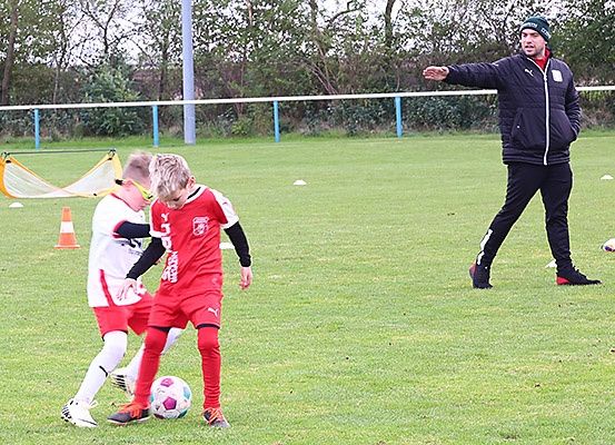 Zwei Jungen spielen Fußball auf einem Feld, einer in Rot und der andere in Weiß. Ein Mann in einer Jacke gibt ihnen Anweisungen. Ein Netz, orange Kegel und Bälle sind auf dem Feld verstreut.
