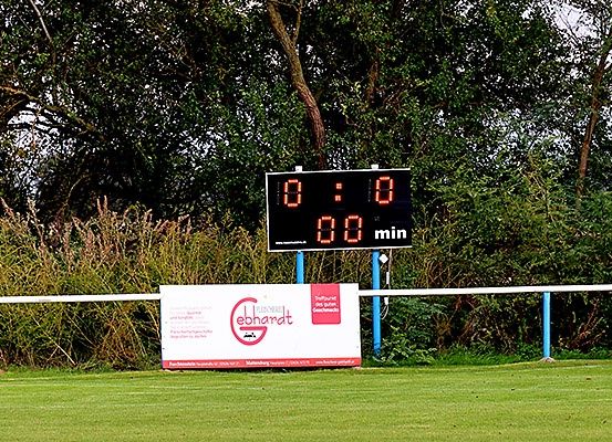 Ein Anzeigetafel zeigt einen Spielstand von 0:0 mit 00 Minuten Spielzeit auf einem Rasenfeld mit Bäumen und Büschen im Hintergrund.
