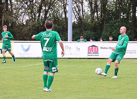Drei Fußballspieler in grünen Trikots sind auf einem Feld. Ein Spieler läuft, während ein anderer den Ball kickt. Der Spieler in der Mitte schaut zurück. Der Hintergrund zeigt eine Zaun mit Werbung und Leuten, die das Spiel beobachten.