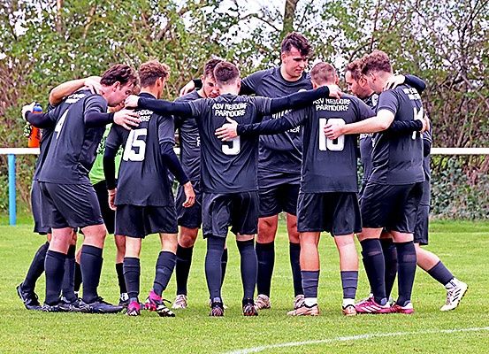 Eine Fußballmannschaft in schwarzen Trikots versammelt sich auf einem Feld, mit Bäumen im Hintergrund, die Nummern 4, 5, 6 und 10 auf den Trikots sichtbar.