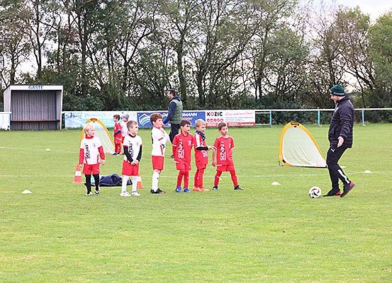 Mehrere junge Kinder in roten Fußballtrikots stehen auf einem Spielfeld und üben mit einem Trainer. Ein Fußballtor befindet sich in der Nähe.