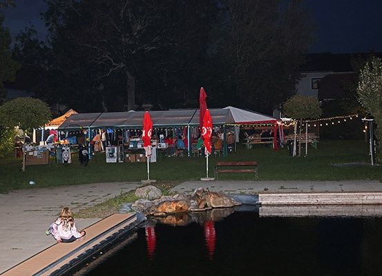 Eine nächtliche Veranstaltung mit einer Outdoor-Aufstellung, einem großen Zelt und einer Frau, die am Teich sitzt. Leute sind unter dem Zelt versammelt.