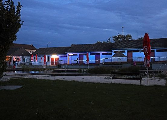 Ein Schwimmbadbereich bei Nacht, beleuchtet von blauen Lichtern. Das Becken ist von einem Zaun umgeben, mit einem Schirm und einer Bank auf dem Rasen.