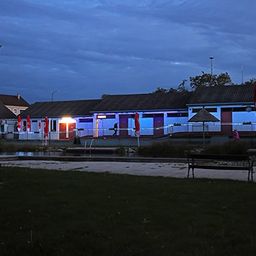 Ein Schwimmbadbereich bei Nacht, beleuchtet von blauen Lichtern. Das Becken ist von einem Zaun umgeben, mit einem Schirm und einer Bank auf dem Rasen.