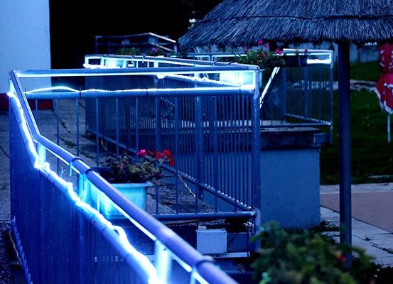 Ein Außenbereich mit einem blauen Metallgeländer, das von blauen Lichtern beleuchtet wird, einem Topf mit roten Blumen und einem strohgedeckten Schirm im Hintergrund.