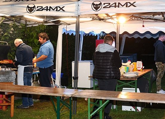 Zwei Männer grillen Essen unter einem Zelt mit 24MX-Marke. Ein Mann in einer Jacke steht in der Nähe eines Tisches mit Gewürzen. Andere Leute sind in der Nähe.