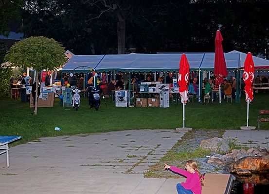 Eine Versammlung von Menschen unter Zelten in einem Außenbereich mit Tischen, Stühlen und einem Tischtennis-Set. Ein Mädchen zeigt auf etwas.
