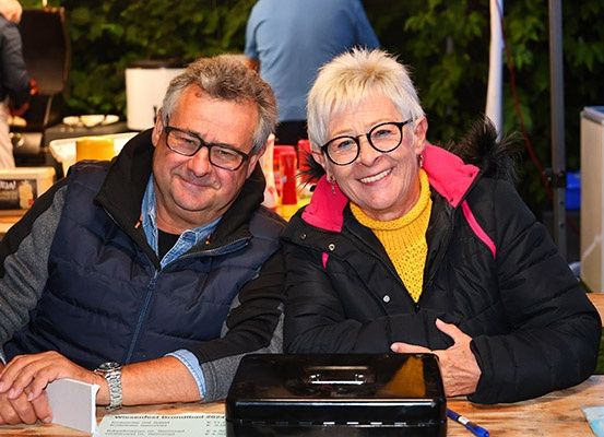 Ein Mann und eine Frau sitzen an einem Tisch und lächeln für ein Foto. Sie tragen beide eine Brille und eine Jacke. Hinter ihnen ist ein Tisch mit verschiedenen Gegenständen aufgebaut.