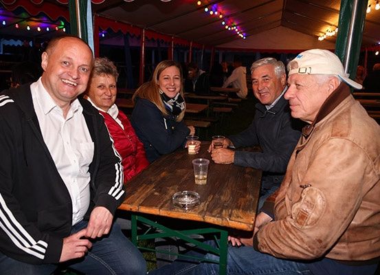 Fünf Menschen lächeln und posieren für ein Foto in einem Außenbereich. Es gibt Tische und Bänke im Bereich, und einige Leute sitzen. Es hängen bunte Lichter von der Decke.