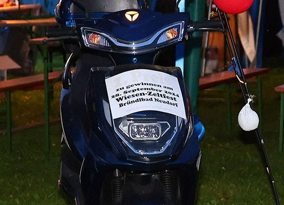 Ein blaues Motorrad mit einem weißen Schild auf der Vorderseite steht im Freien. Das Schild trägt die Aufschrift 'Zu gewonnen am 28. September 2024 Wiesbaden-Zeltfest Brundbad Neudorf'. Im Hintergrund befinden sich Tische und Bänke.