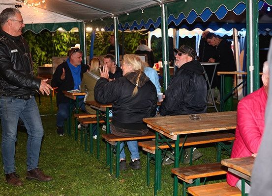Eine Gruppe von Menschen sitzt an Tischen unter einem Zelt. Ein Mann redet und gestikuliert, während die anderen zuhören und lachen.
