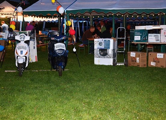 Zwei Roller sind auf einer Wiese geparkt, in der Nähe eines Zelts mit Luftballons. Unter dem Zelt sitzen Menschen an Tischen, und in der Nähe befinden sich Kisten und eine Waschmaschine.