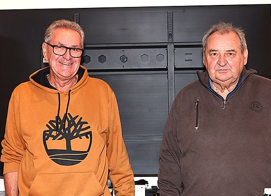 Zwei ältere Männer stehen nebeneinander und lächeln vor einem schwarzen Metallgerüst.