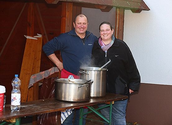 Ein Mann und eine Frau stehen lächelnd neben großen Töpfen auf einem Holztisch. Sie scheinen zu kochen.