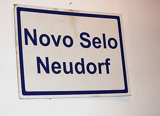 Ein weißes Schild mit blauen Rändern an einer Wand montiert. Es steht 'Novo Selo Neudorf' in fetten, blauen Buchstaben.