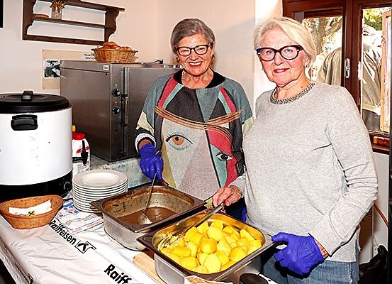 Zwei Frauen in Brillen und Handschuhen stehen neben einem Buffettisch mit Essen in einer Küche. Eine hält einen Kochlöffel.