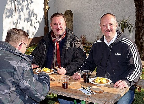 Drei Männer sitzen an einem Tisch und essen und trinken. Der Mann in der Mitte hat einen Teller mit Essen und ein Getränk. Der Mann rechts trägt eine Adidas-Jacke.