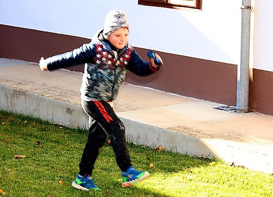 Ein junger Junge läuft auf einem Grasfeld und trägt eine Winterjacke und Handschuhe. Er hat einen blauen Gegenstand in der Hand. Hinter ihm steht ein Gebäude mit einer weißen Wand und einem braunen Rand.