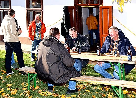 Eine Gruppe von Männern sitzt an einem Picknicktisch auf einer Rasenfläche, einige mit Getränken in der Hand. Hinter ihnen stehen zwei Männer und einer hält eine Flasche. Ein Gebäude ist im Hintergrund.
