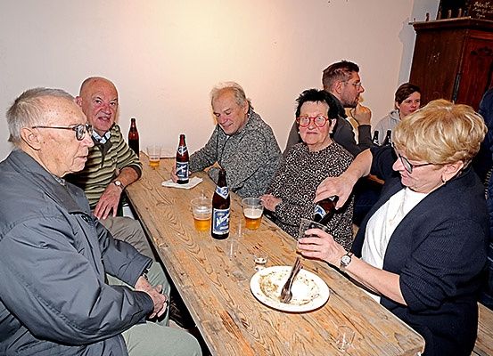Eine Gruppe älterer Personen, sowohl Männer als auch Frauen, sitzen um einen Holztisch herum, möglicherweise um eine Mahlzeit oder Getränke zu genießen. Sie haben verschiedene Getränke auf dem Tisch, darunter Bierflaschen und Gläser.