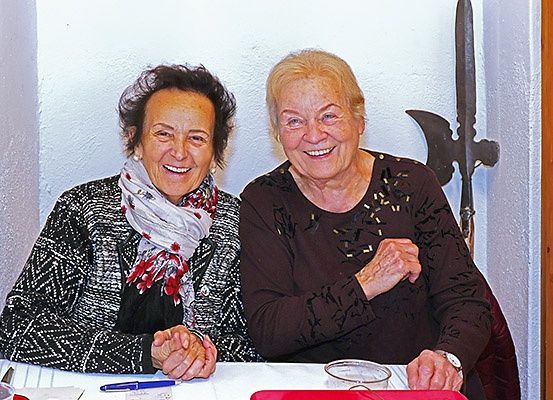 Zwei lächelnde Frauen sitzen an einem Tisch, eine in einer schwarz-weißen Jacke und die andere in einem braunen Oberteil, beide scheinen für ein Foto zu posieren. Hinter ihnen lehnt eine Lanze an einer weißen Wand.