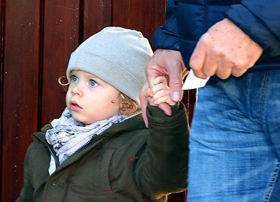 Ein junges Kind mit blauen Augen, das einen grünen Mantel, eine Mütze und einen Schal trägt, hält ein Stück Papier in der Hand, während es neben einem erwachsenen Mann in blauer Jeans steht.