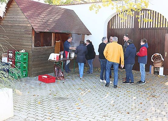 Eine Gruppe von Menschen hat sich vor einem Holzschuppen versammelt, wo ein Mann auf einem Herd kocht. Es stehen mehrere Menschen herum und beobachten.