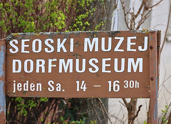 Ein hölzerne Schild zeigt 'SEOSKI MUZEJ DORFMUSEE'. Darunter steht in kleineren Buchstaben 'jeden Sa. 14 - 16 30h'. Das Schild ist verwittert und in einer natürlichen Umgebung platziert.