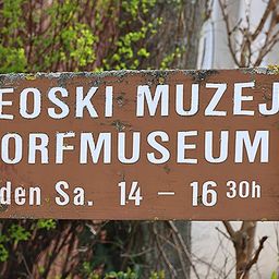 Ein hölzerne Schild zeigt 'SEOSKI MUZEJ DORFMUSEE'. Darunter steht in kleineren Buchstaben 'jeden Sa. 14 - 16 30h'. Das Schild ist verwittert und in einer natürlichen Umgebung platziert.