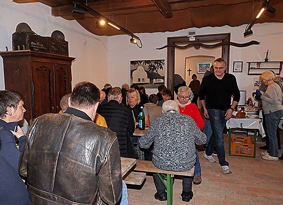 Eine Gruppe von Menschen versammelt sich in einem Raum mit Ziegelboden. Einige sitzen auf Bänken und andere stehen um einen Tisch herum. Ein Mann in einem schwarzen Shirt geht in der Nähe des Tisches, und eine Frau in einem roten Shirt sitzt. Ein Schrank und Bilderrahmen sind an der Wand montiert.