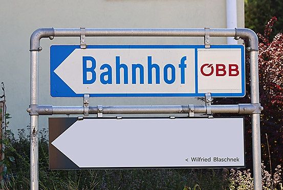 Ein blau-weißes Schild mit der Aufschrift 'Bahnhof' und einem Pfeil nach links. Darunter befindet sich ein weißes Schild mit der Aufschrift 'Wilfried Blaschnek'. Das Schild ist an einem Metallpfosten befestigt.