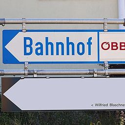 Ein blau-weißes Schild mit der Aufschrift 'Bahnhof' und einem Pfeil nach links. Darunter befindet sich ein weißes Schild mit der Aufschrift 'Wilfried Blaschnek'. Das Schild ist an einem Metallpfosten befestigt.