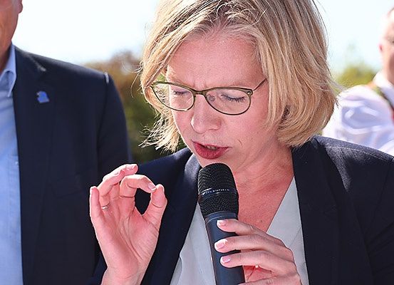 Eine Frau mit Brille spricht in ein Mikrofon, während ein Mann in Anzug im Hintergrund steht.