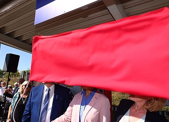 Drei Personen stehen unter einem überdachten Bereich, zwei halten eine rote Fahne. Ein Lautsprecher steht in der Nähe mit einer Band im Hintergrund.